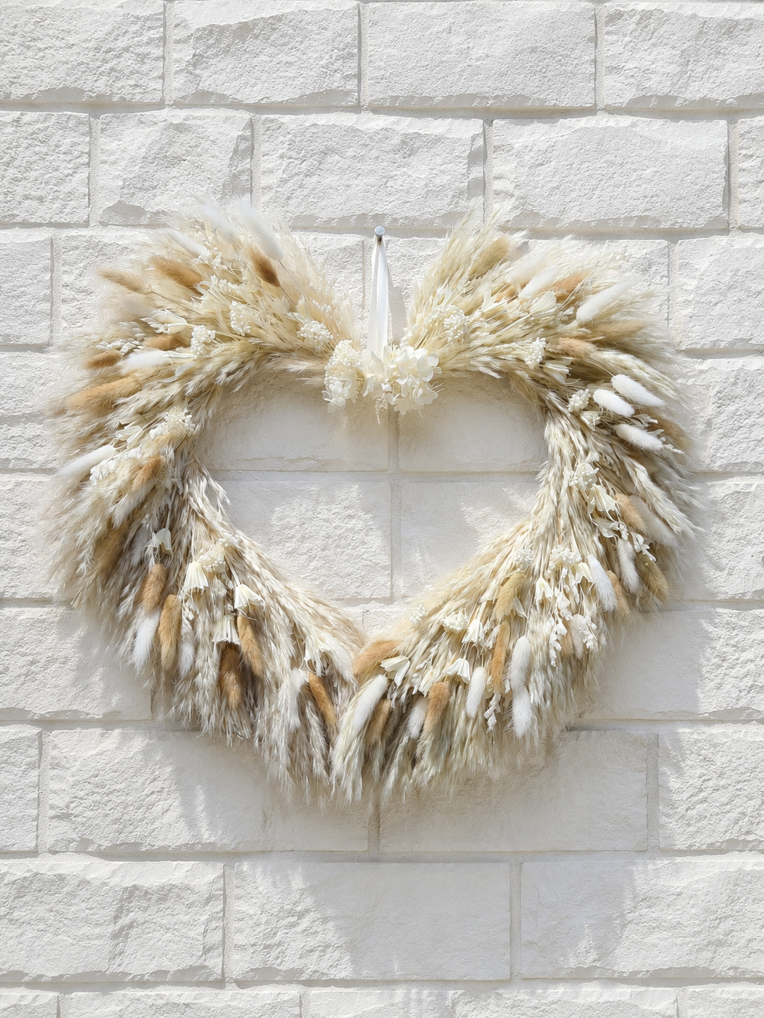 Heart-shaped dried pampas grass wreath in beige and white hanging on a white brick wall.