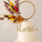 A two-tier cream cake on a light wood stand with a gold ring topper and dried floral accents.