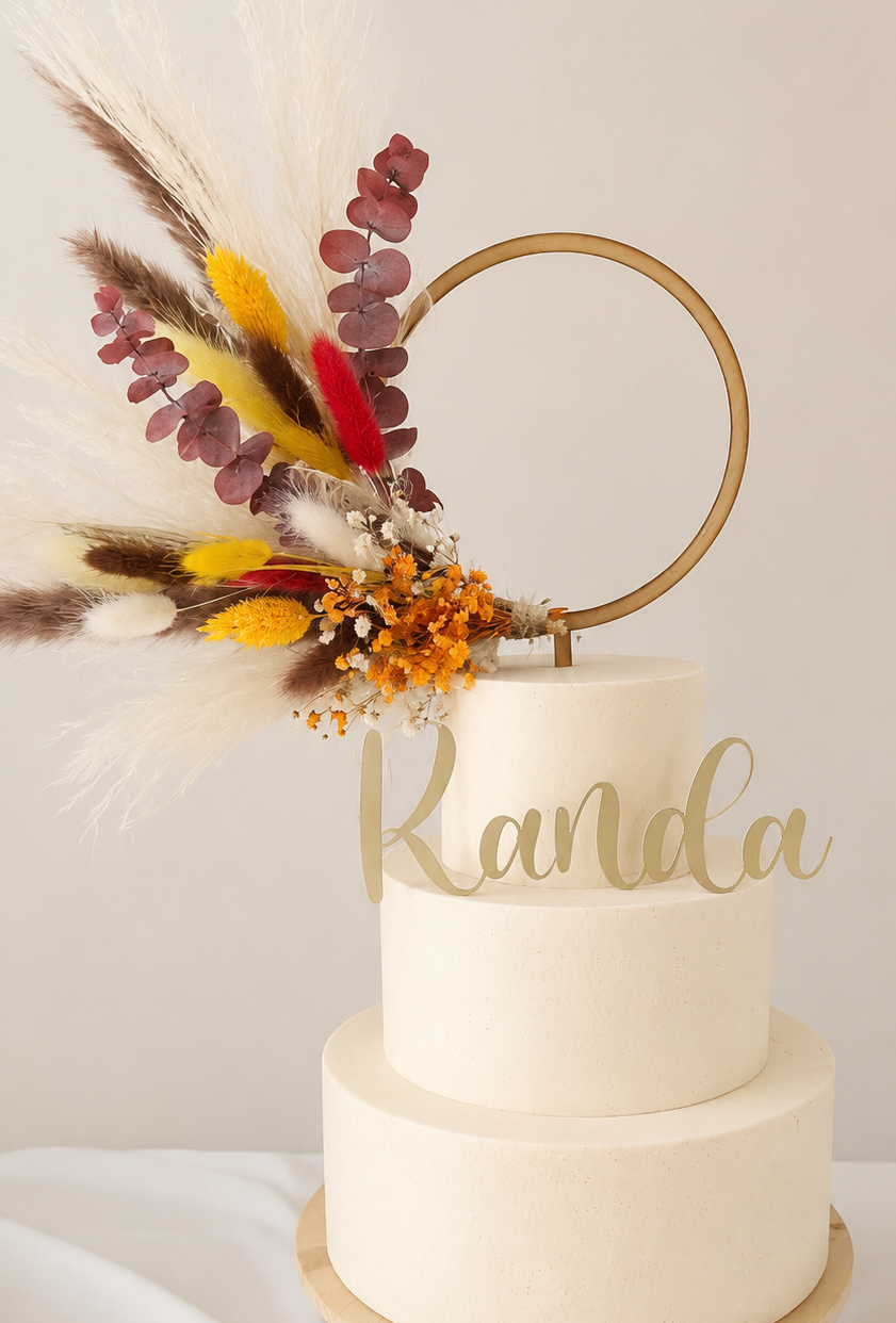 A two-tier cream cake on a light wood stand with a gold ring topper and dried floral accents.