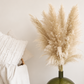 A glossy olive green glass vase holding tall natral pampas grass, styled on a light surface beside a white textured pillow and draped white fabric.