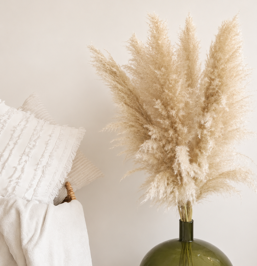 A glossy olive green glass vase holding tall natral pampas grass, styled on a light surface beside a white textured pillow and draped white fabric.