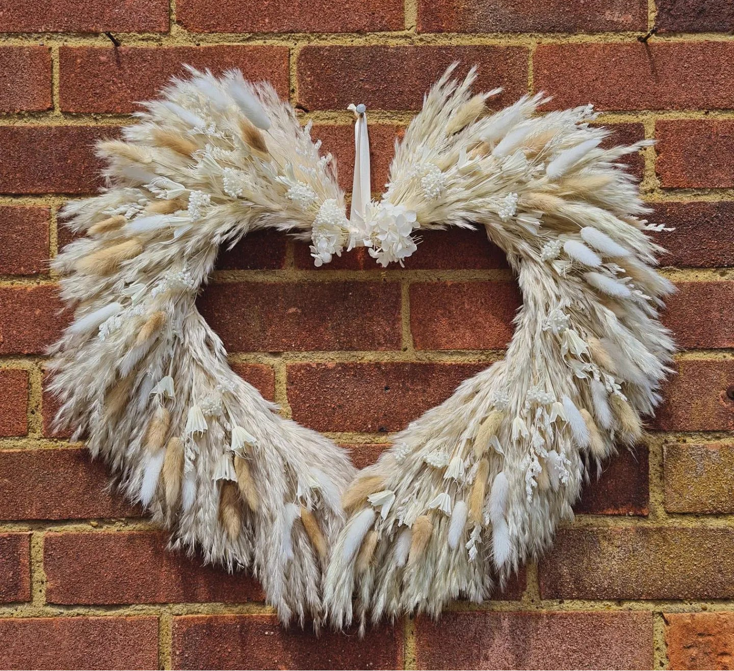 A heart-shaped dried pampas grass wreath hanging on a red brick wall.
